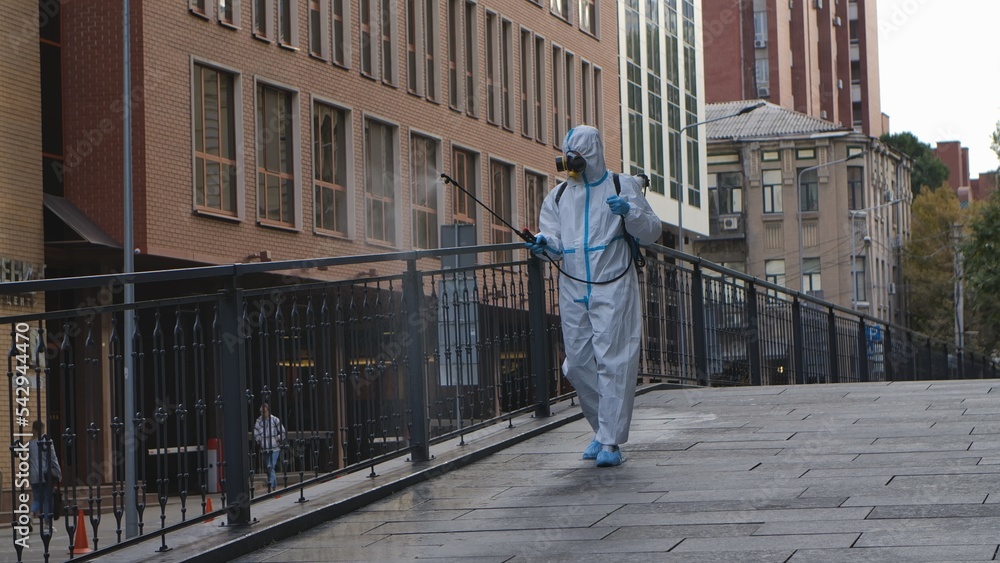 Medical worker in white protective suit and respirator disinfects railing on city street. Virologist treating objects with an antiseptic using an alcohol disinfectant and sprinkler.