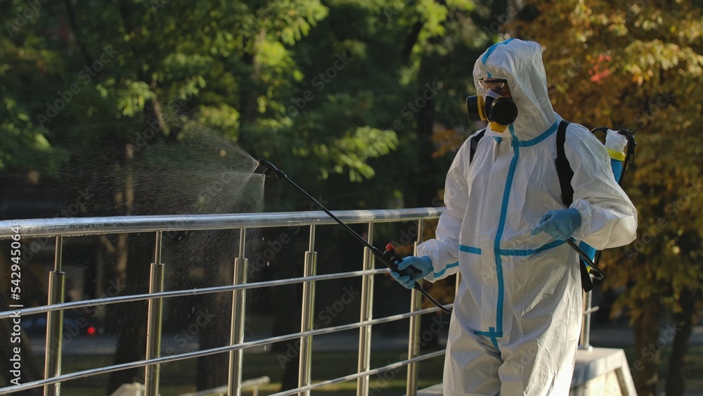A virologist in a white protective suit and respirator disinfects railings on city streets. Disinfection of objects with antiseptic spray to prevent the spread of the Covid 19 outbreak.