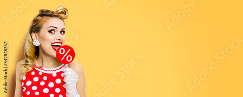 Very tasty discounts, rebates, deals, sales concept. Cheerful beautiful woman licking signboard with % sign, dressed in pinup red dress, over yellow background. Happy girl at studio. Wide picture