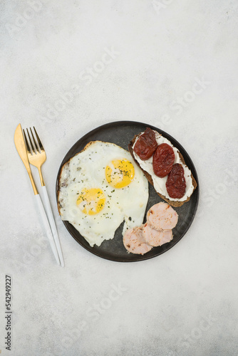 Balanced breakfast of two fried eggs, sandwich of cream cheese and dried tomatoes, slices of poultry meat on gray background.
