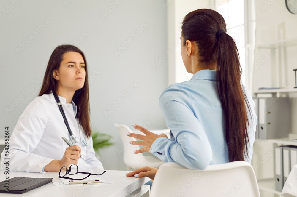 Young professional Caucasian woman in doctor coat looks at female patient of clinic during medical consultation on health problems sits with back to camera, posing at table in therapist office Young professional Caucasian woman in doctor coat looks at female patient of clinic during medical consultation on health problems sits with back to camera, posing at table in therapist office