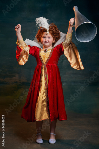 Portrait of little red-headed girl  child in costume of royal person isolated over dark green background. Winning look