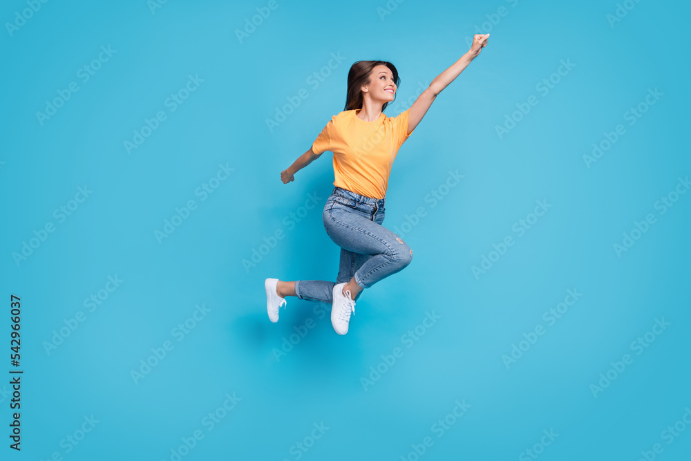 Full length photo of excited cool lady wear yellow t-shirt jumping high rising fist isolated blue color background Full length photo of excited cool lady wear yellow t-shirt jumping high rising fist isolated blue color background
