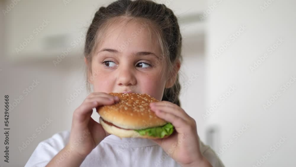 little girl eating a hamburger. unhealthy fast food proper nutrition concept. child greedily with lifestyle pleasure bites a big burger in the kitchen at home. kid eats fast food close-up little girl eating a hamburger. unhealthy fast food proper nutrition concept. child greedily with lifestyle pleasure bites a big burger in the kitchen at home. kid eats fast food close-up