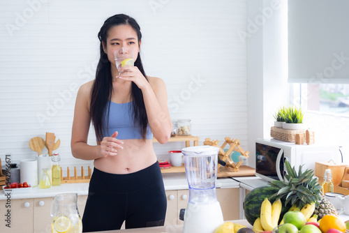 Fit and healthy young Asian woman in sportswear preparing and drinking fresh lemon water for healthy lifestyle and fitness in modern kitchen after morning workout and exercise