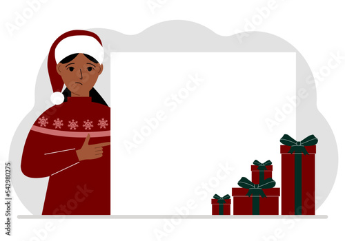 A woman in a Christmas or New Year's sweater with a red sana cap points his finger at a banner, a blank poster or a large white sheet of paper with space for text.