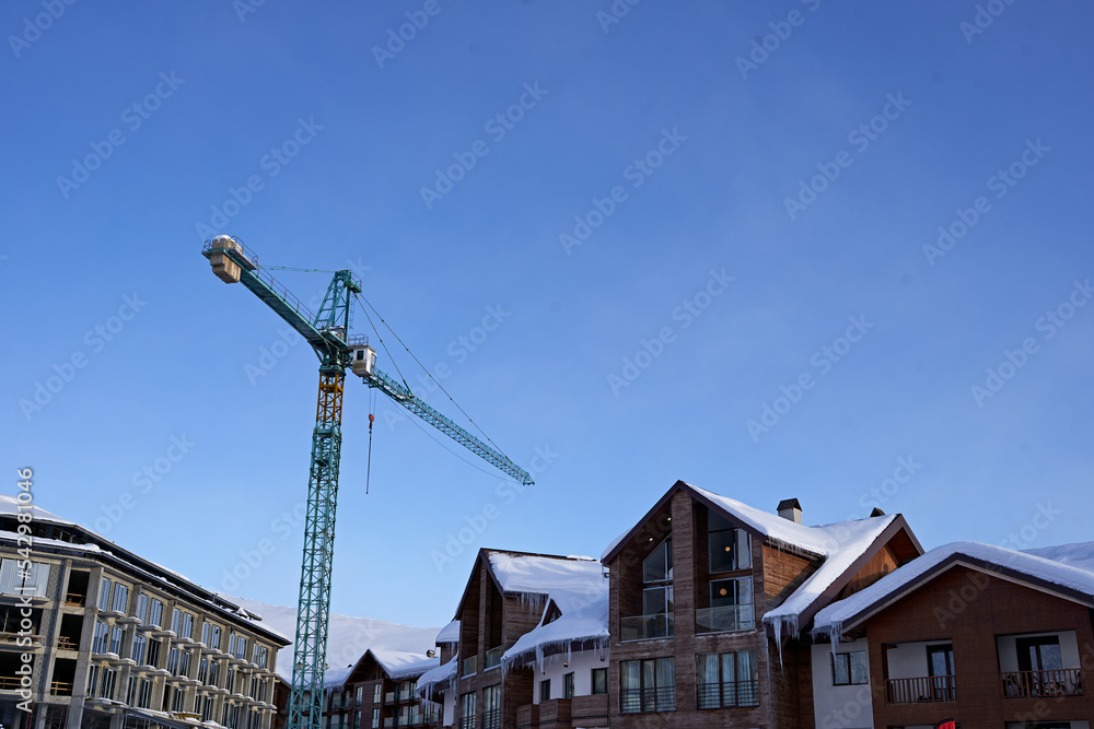 Construction site of ski resort hotel chalet framework and crane with snowy mountain ridge on winter sunrise on background. Mountains range covered with snow powder against the residental building.