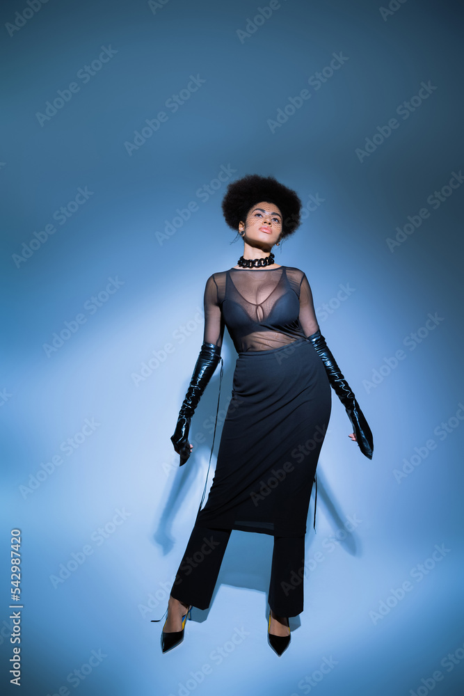 full length of curly african american woman in transparent shirt and black skirt looking up on blue.