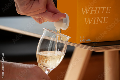 Hand pouring white wine into a glass from a BIB - cardboard bag in box with open tap standing on a table. Close up image. Fictitious brand.