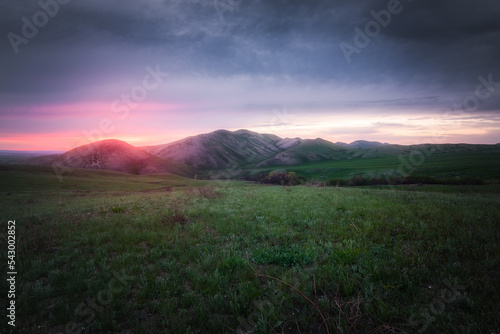 View of the hills and mountains covered by green grass under purple cloudy sky lightened by rising sun