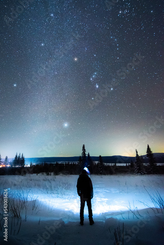 A man with a headlamp stands at night on a winter path under a bright starry sky and the Orion constellation