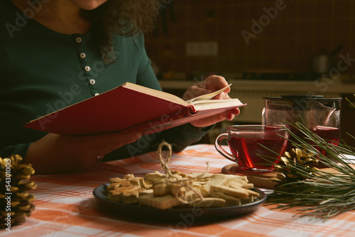 Cropped view of a woman in green shirt, reading harcover book or novel while a tea time with hibiscus hot drink and Christmas gingerbread cookies. Enjoy winter holidays at home. Festive mood. New Year photo