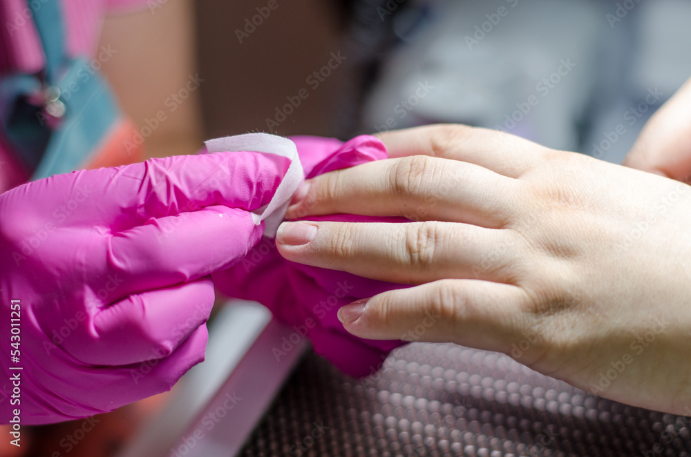 Female manicurist in pink gloves works with young hands and nails under a bright lamp. Process of wiping the nail with a lint-free cloth from dust