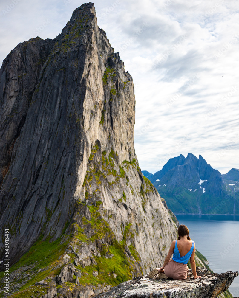 Fototapeta premium a beautiful girl sits on the rocks enjoying the view of the famous segla mountain in norway, senja island; hesten trail head, the famous norwegian fjords with mighty mountains above the sea