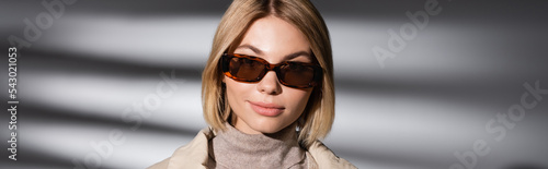Stylish woman in warm turtleneck and sunglasses standing on abstract grey background, banner.