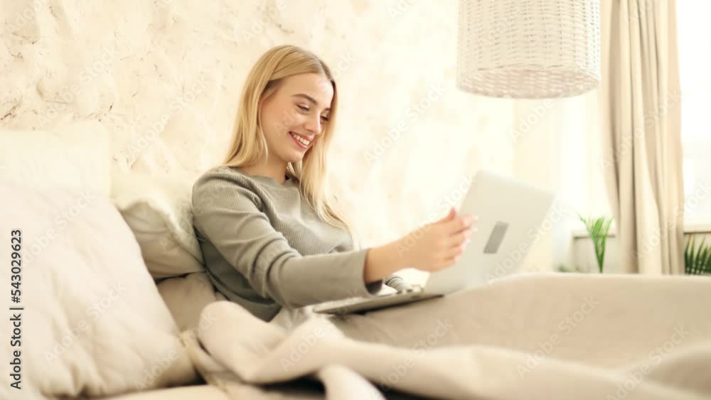 Young attractive blond woman have a video call while laying on a bed in light living room. Young lady talking on video conference online indoors. Young attractive blond woman have a video call while laying on a bed in light living room. Young lady talking on video conference online indoors.