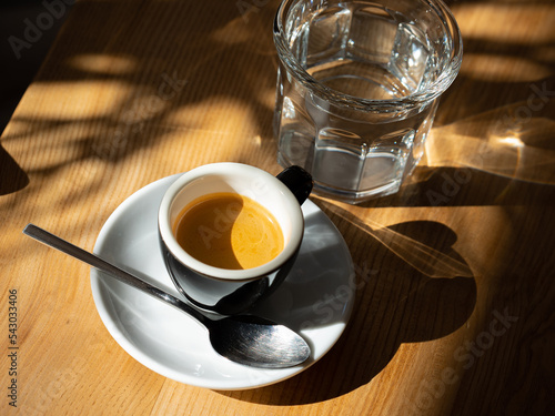 Espresso in a black cup served with glass of water on wooden table under sunlight