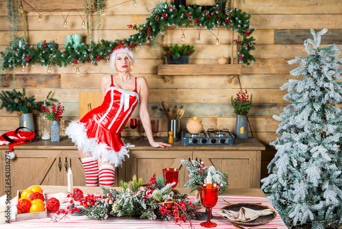 slim blond woman wearing red xmas gown, hat and striped half hose is standing in the wooden kitchen and cooking breackfast photo