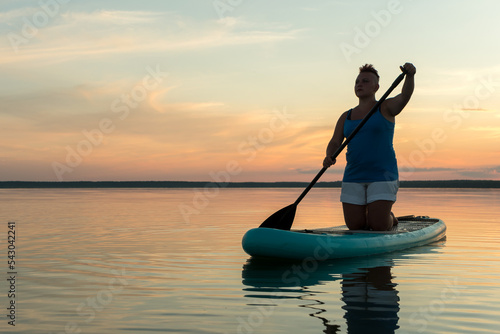 A woman with a Mohawk in shorts on her knees on a SUP board with an oar against the backdrop of a bright sunset sky swims in the lake in the evening.