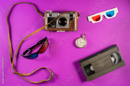 Silver vintage pocket watch and 3D glasses with blue red stereoscopic lenses. Black plastic VHS video cassette and retro camera on purple background. Retro, vintage, history, nostalgia concept.