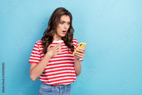 Photo of young funny nervous unexpected woman wear red striped t-shirt touching chest hold phone looking her facebook account isolated on blue color background