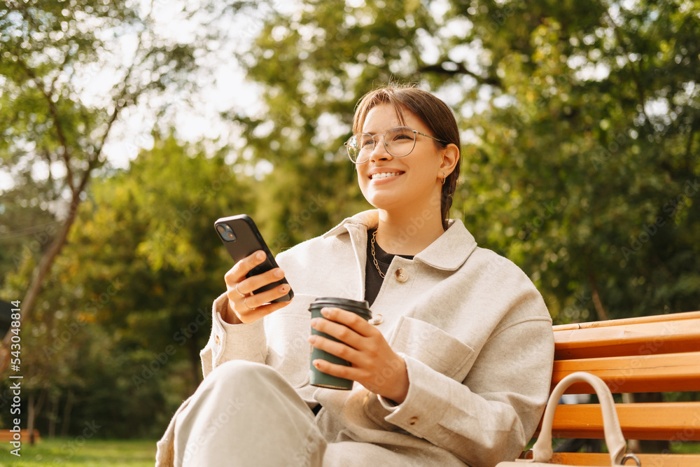 Portrait of smiling young woman wearing glasses and looking away and holding smartphone and cup of coffee. Portrait of smiling young woman wearing glasses and looking away and holding smartphone and cup of coffee.