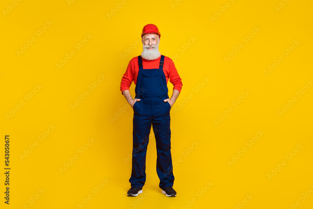Full length photo of good mood senior man builder with white beard red helmet hands in pockets isolated on yellow color background Full length photo of good mood senior man builder with white beard red helmet hands in pockets isolated on yellow color background