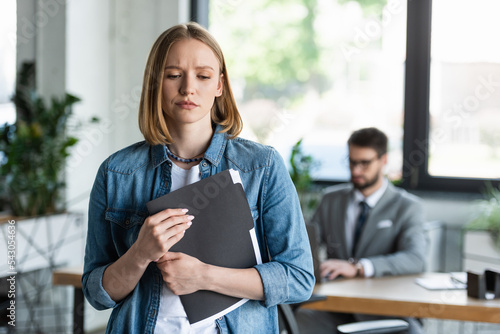 Sad woman holding paper folder with resume in blurred office.