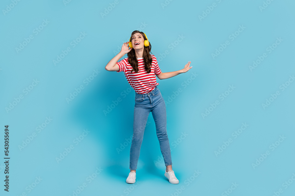 Full length photo of funky dreamy lady wear striped t-shirt headphones having fun singing isolated blue color background Full length photo of funky dreamy lady wear striped t-shirt headphones having fun singing isolated blue color background