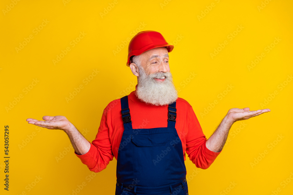 Photo of cheerful positive age man workwear overall red hard hat looking comparing arms empty space isolated yellow color background Photo of cheerful positive age man workwear overall red hard hat looking comparing arms empty space isolated yellow color background
