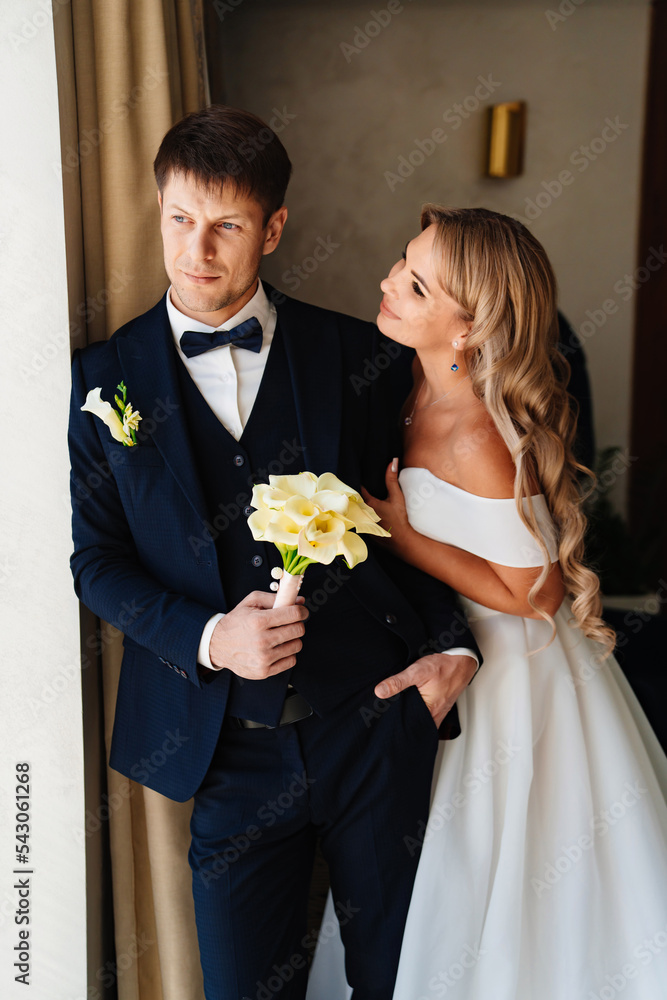 the blonde bride gently hugs the groom's shoulders by the window. the blonde bride gently hugs the groom's shoulders by the window.