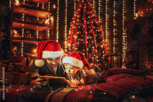Dad with little son in pajamas and Santa's hats lying in bed uses digital tablet before sleeping