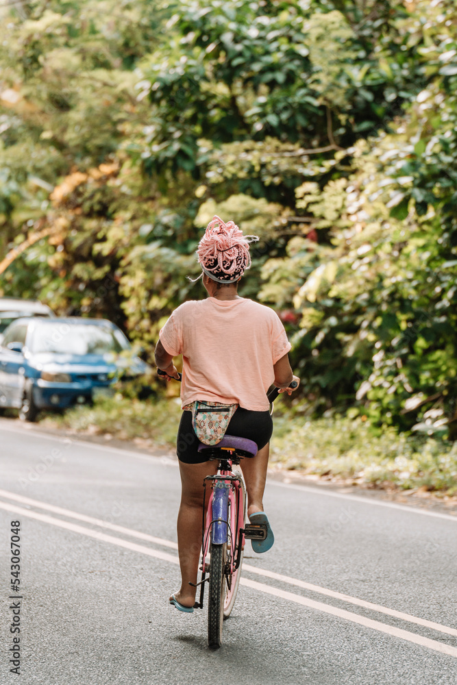 Imagen vertical desde atrás de una joven mujer afroamericana con cabello rosado andando en bicicleta por la calle en un día de verano. 