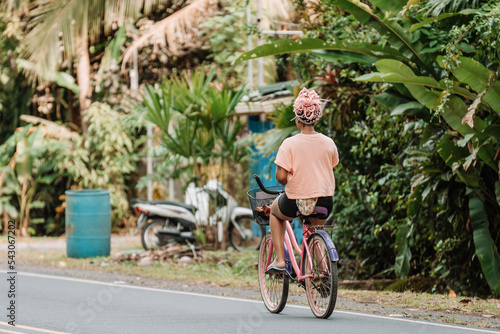 Imagen horizontal desde atrás de una joven mujer afroamericana con cabello rosado andando en bicicleta por la calle en un día de verano. 
