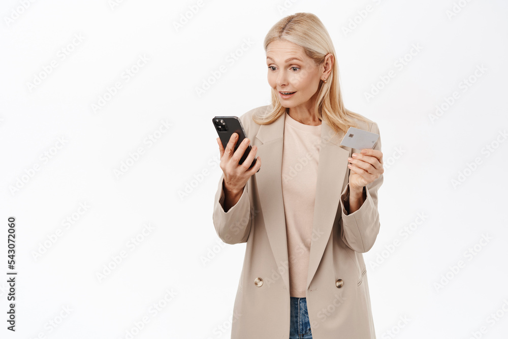 Online shopping. Smiling blond elegant woman looking at mobile phone, holding credit card, paying in application, standing over white background Online shopping. Smiling blond elegant woman looking at mobile phone, holding credit card, paying in application, standing over white background