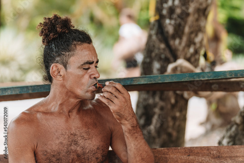 Imagen horizontal de un hombre adulto latino con cabello rizado sin camisa en la playa fumando hierba con una pipa en un día de verano. 
