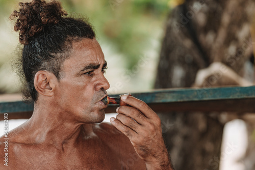 Imagen horizontal de un hombre adulto latino con cabello rizado sin camisa en la playa fumando hierba con una pipa en un día de verano. 