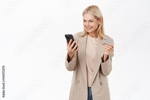 Online shopping. Smiling blond elegant woman looking at mobile phone, holding credit card, paying in application, standing over white background