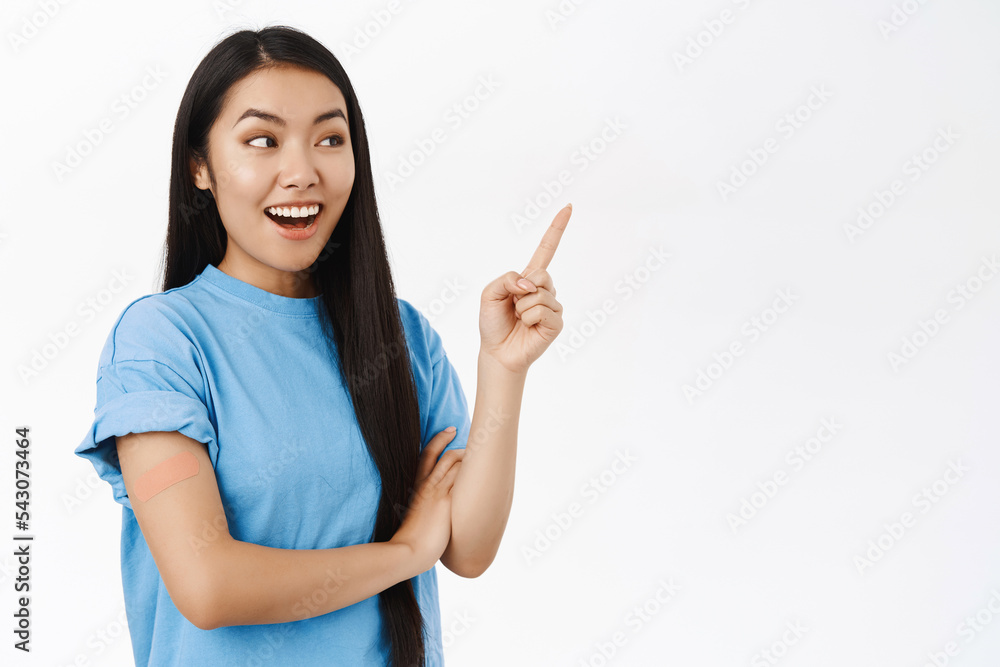 Enthusiastic asian girl got vaccinated, has band aid patch on shoulder after covid vaccine, smiling and pointing at copy space banner, showing coronavirus info announcement Enthusiastic asian girl got vaccinated, has band aid patch on shoulder after covid vaccine, smiling and pointing at copy space banner, showing coronavirus info announcement