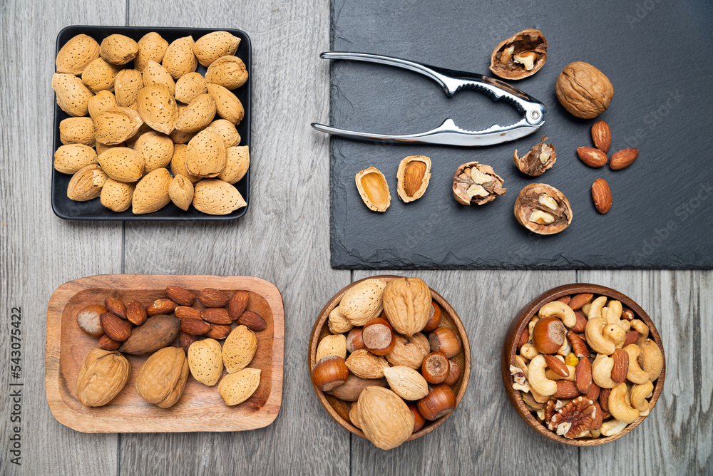 nuts in bowls and a nutcracker on a gray table. top view, Assortment of nuts - cashew, hazelnuts, almonds, walnuts, pistachio, pecans, pine nuts, peanut. nuts in bowls and a nutcracker on a gray table. top view, Assortment of nuts - cashew, hazelnuts, almonds, walnuts, pistachio, pecans, pine nuts, peanut.