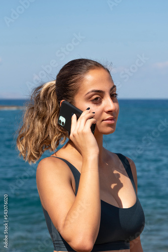 Malia, Crete, Greece. 2022. Portrait of an ttractive woman with ponytail hair using a mobile phone at the seaside.