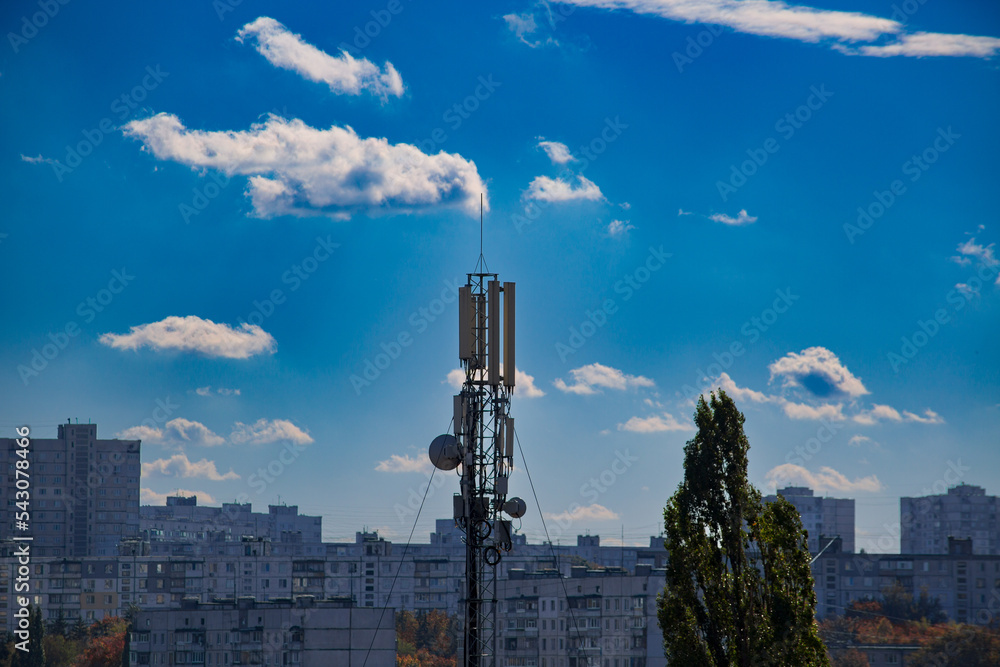 Fototapeta premium Mobile communication tower in the city among the houses against the blue cloudy sky. Internet connection in the big city 5g and 4g. Mobile communication in the city