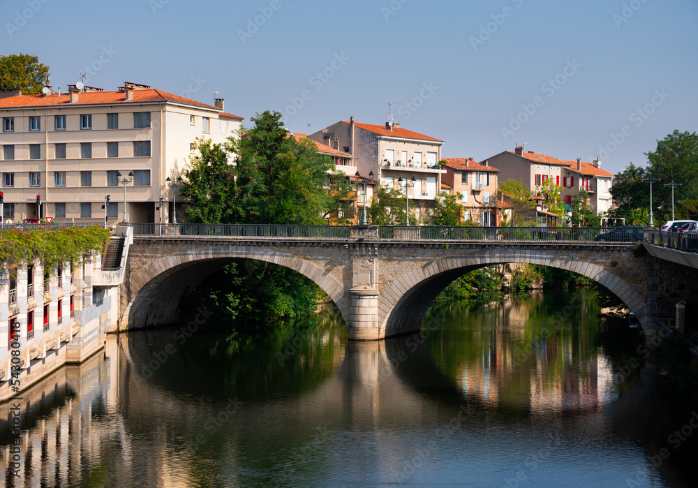 Scenic view of summer townscape of Castres with modern townhouses with terracotta tiled roofs on bank of Agout river and old Pont Miredames arched bridge on sunny day, France