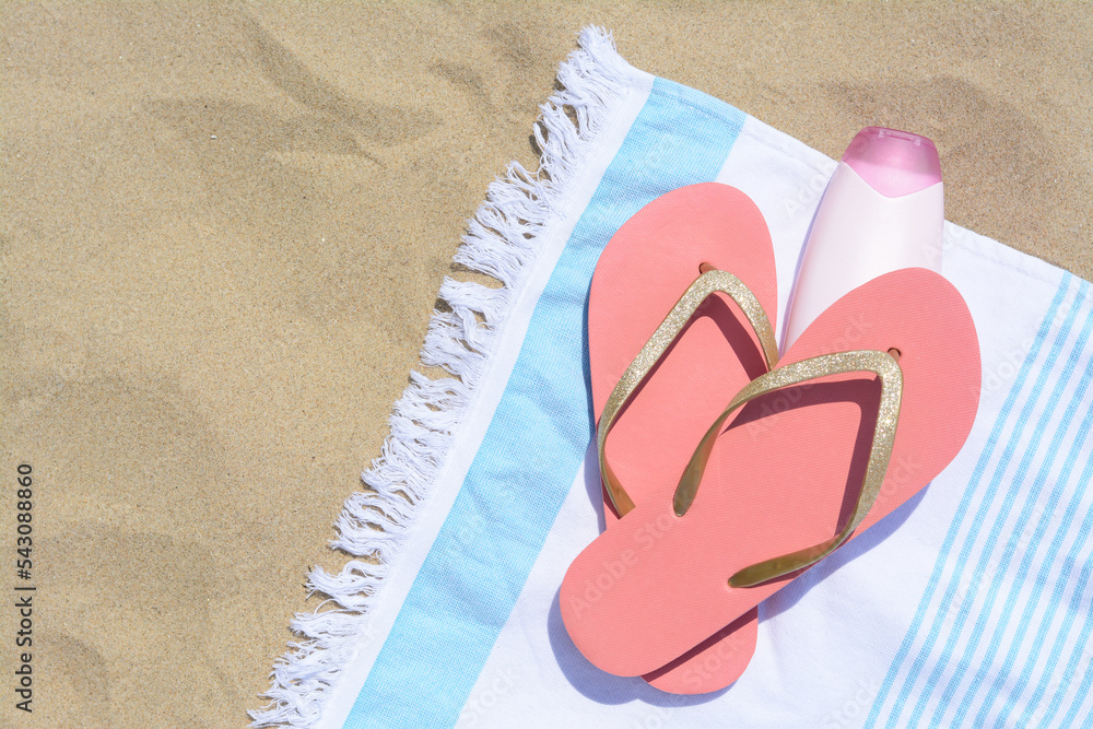 Striped towel with bottle of sunblock and flip flops on sandy beach, flat lay. Space for text Striped towel with bottle of sunblock and flip flops on sandy beach, flat lay. Space for text