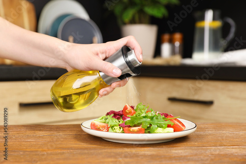 Woman spraying cooking oil onto salad on wooden table in kitchen, closeup