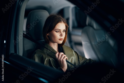 a sweet, relaxed, stylish woman is sitting in a black car in the passenger seat, at night and wearing a seat belt. Topics of road safety © SHOTPRIME STUDIO