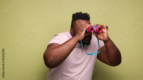 Bearded attentive concentrated young adult African-american man wearing pink t-shirt stranding with binoculars, spying. Indoor studio shot isolated on light green background.