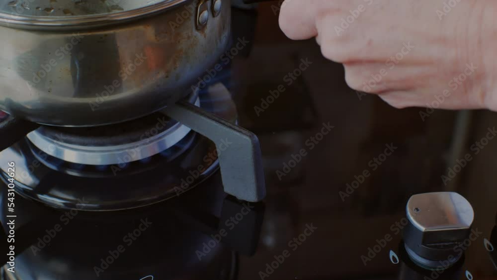 Close-up of a hand igniting gas by turning the handle of a gas stove and placing a stainless steel saucepan with a shiny reflection on a burning burner. Modern gas stove in the kitchen. Home cooking Close-up of a hand igniting gas by turning the handle of a gas stove and placing a stainless steel saucepan with a shiny reflection on a burning burner. Modern gas stove in the kitchen. Home cooking
