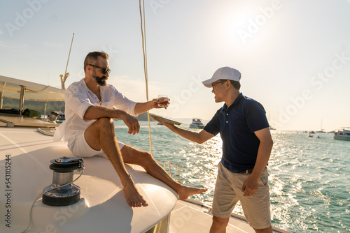 Asian man waiter serving alcohol drink to passenger tourist while catamaran boat yacht sailing in the ocean at summer sunset. Handsome man enjoy luxury outdoor lifestyle on holiday travel vacation. © CandyRetriever 
