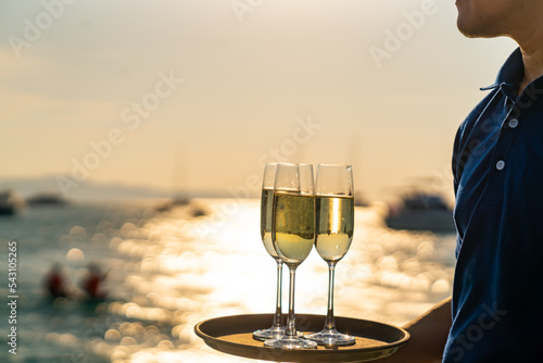 Asian man waiter holding champagne glass on the tray serving to group of passenger tourist travel on luxury catamaran boat yacht sailing in the ocean at summer sunset on beach holiday vacation trip.